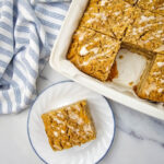 apple crumb cake in a pan with one slice cut out and placed on a white plate.