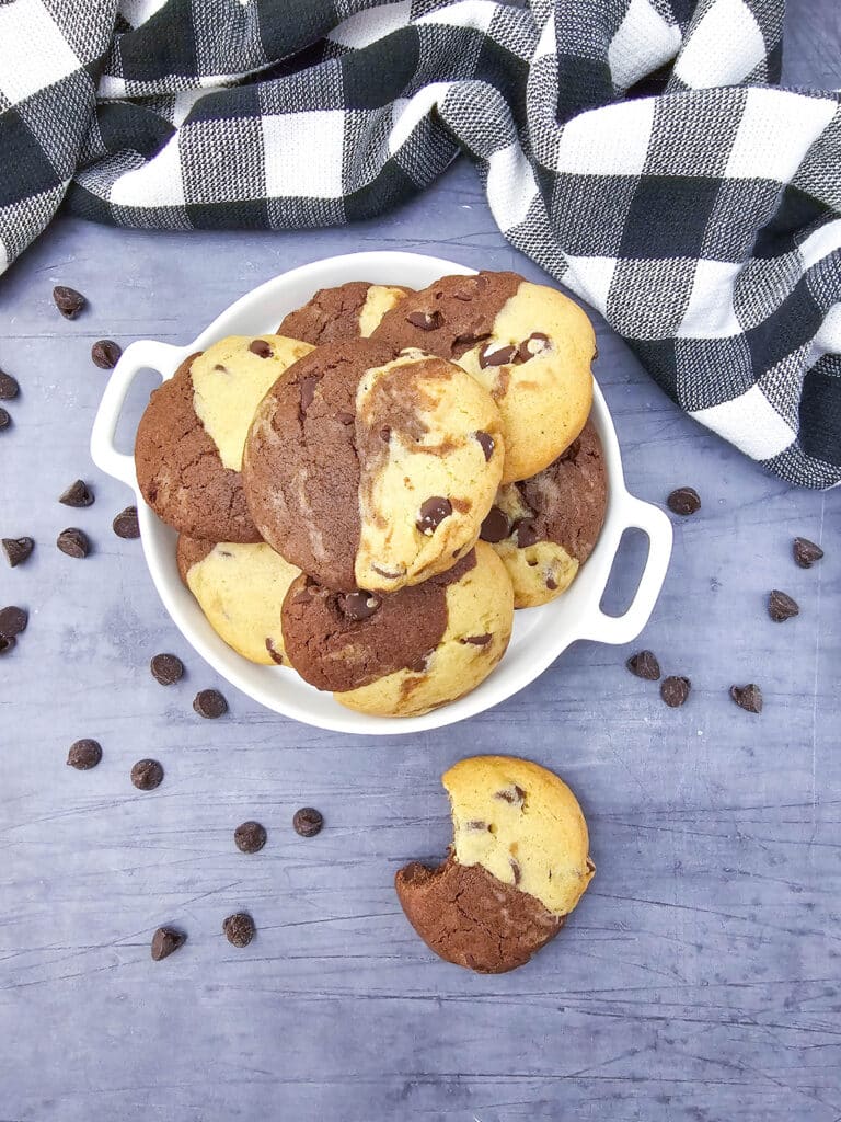 Top-down view of a white handled dish filled with brookies and a bitten cookie on a gray wood surface surrounded by scattered chocolate chips.