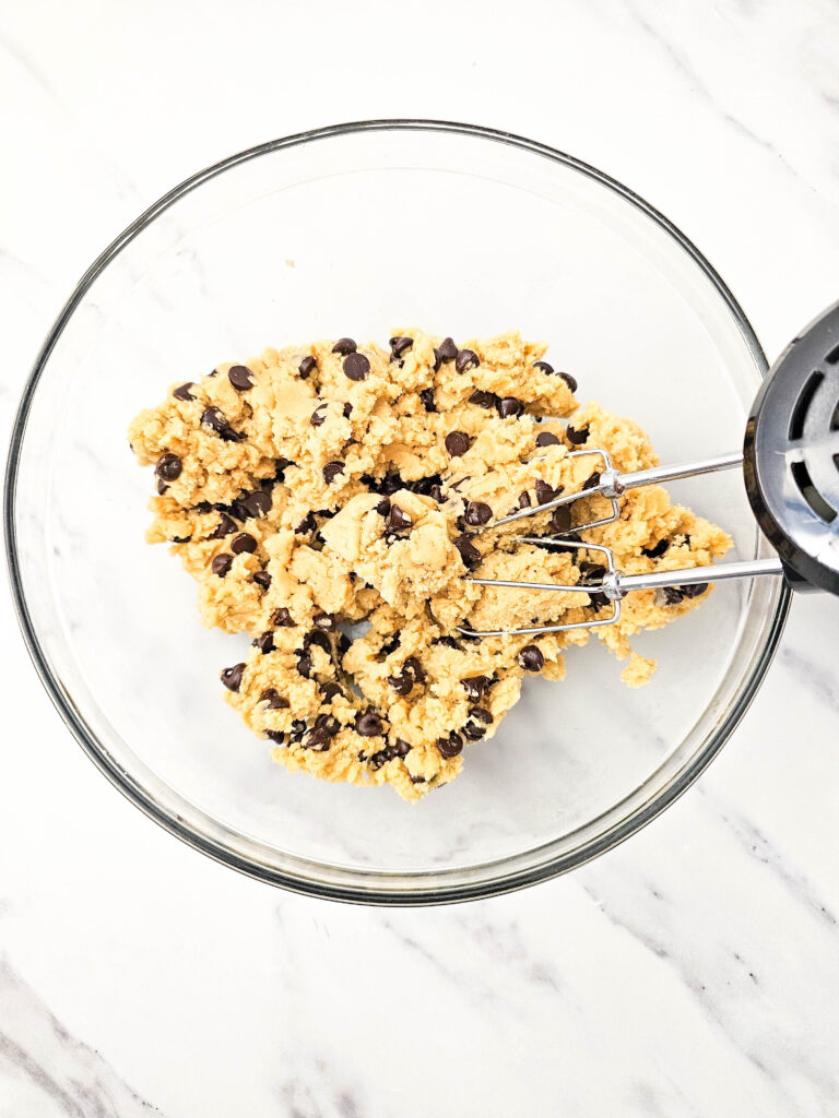 Cookie dough half with chocolate chips stirred in, showing the golden brookies recipe dough in a glass bowl.