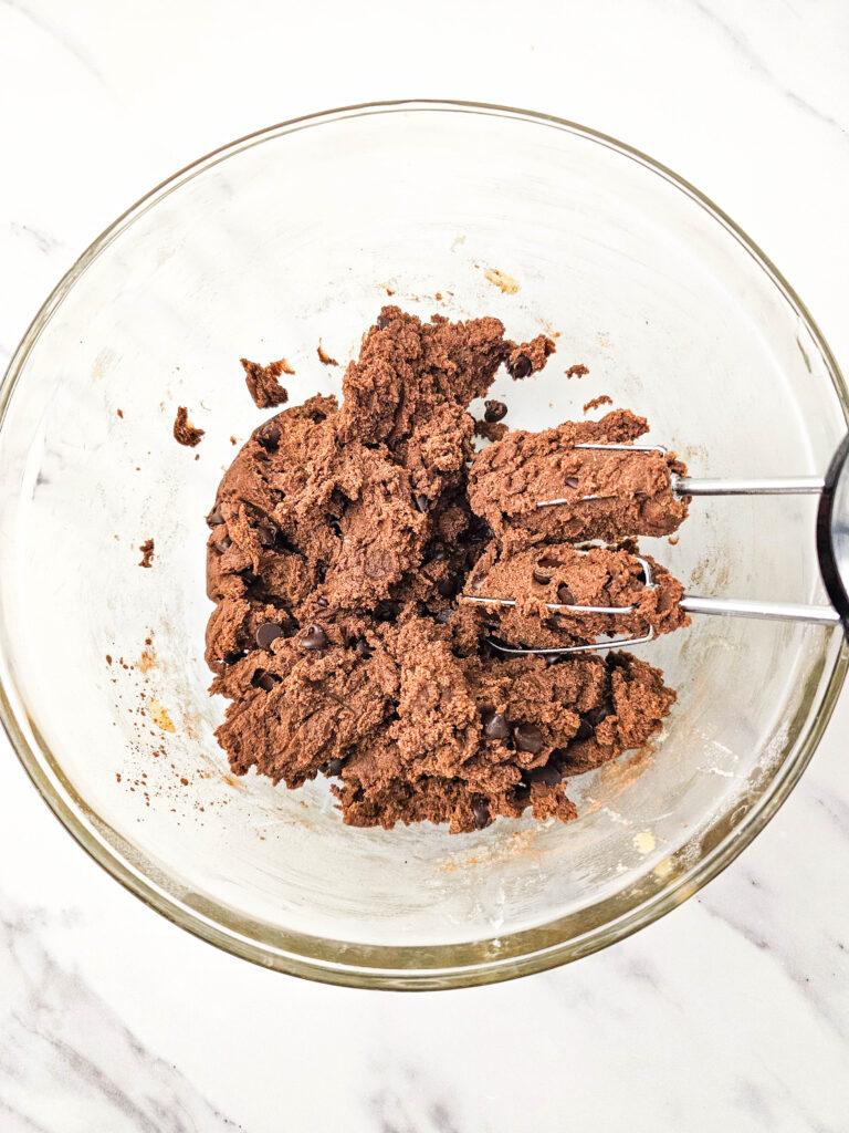 Finished chocolate brownie dough with chocolate chips fully mixed in, shown in a glass bowl with a hand mixer.