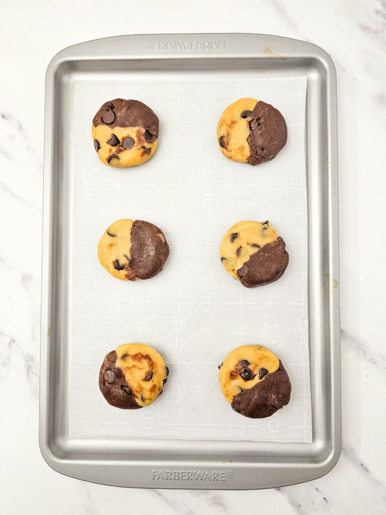 Six brookies recipe dough portions on a parchment-lined baking sheet, each showing the brownie and cookie dough halves pressed together and slightly flattened.