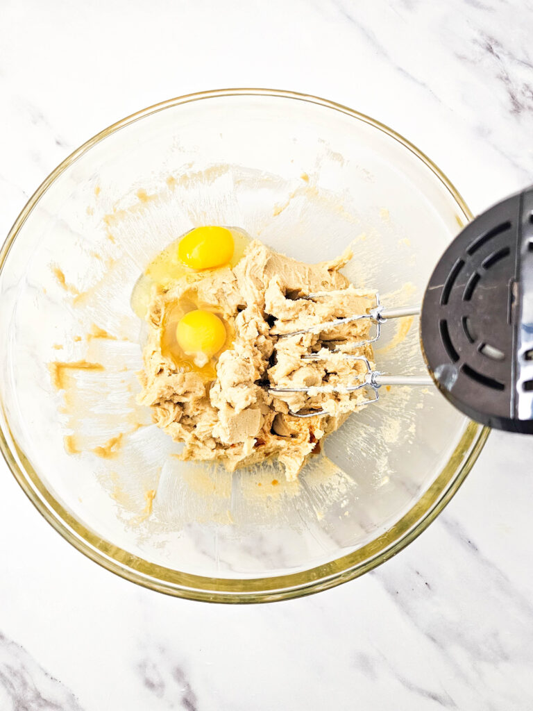 Two eggs cracked into creamed butter and sugar mixture in a glass bowl with a hand mixer.