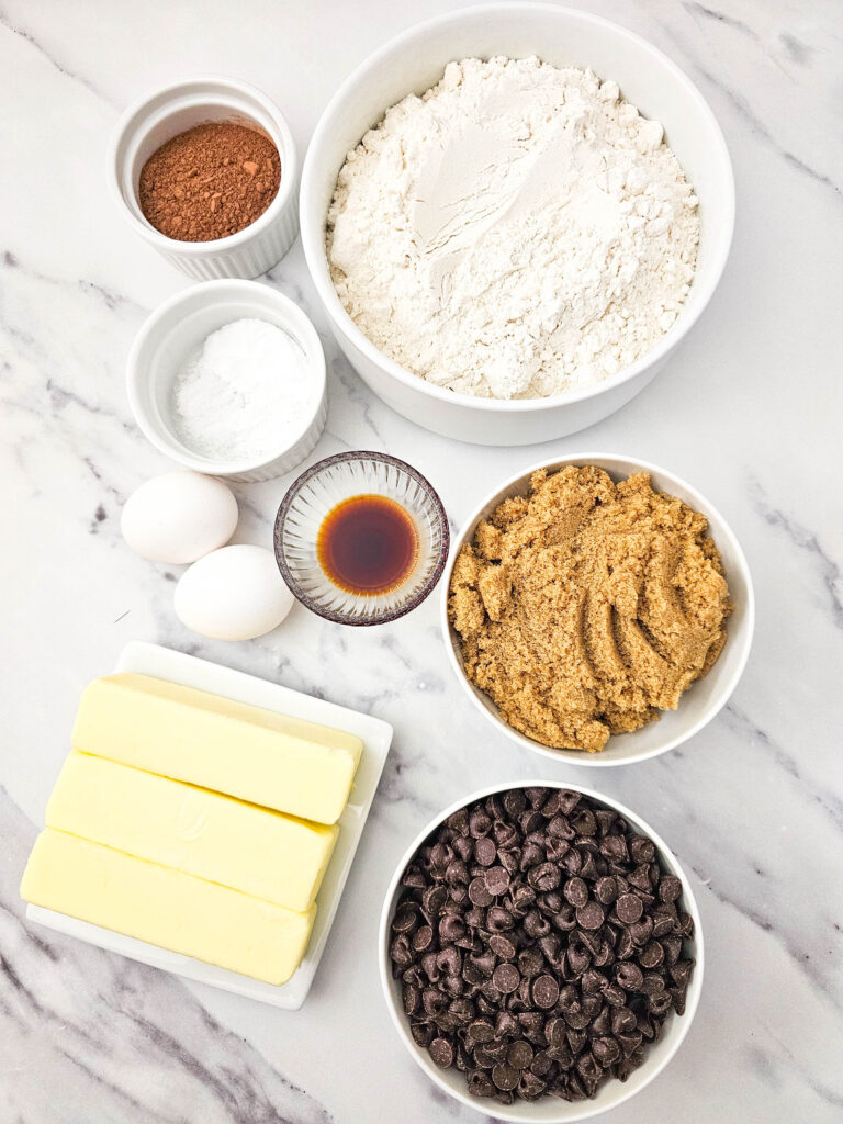 Flat lay of brookies recipe ingredients on a marble surface including three sticks of butter, brown sugar, eggs, vanilla, flour, cocoa powder, and semi-sweet chocolate chips.