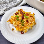 Serving of Million Dollar Potato Casserole on a white plate with buttery cracker topping, bacon pieces, and green onions.