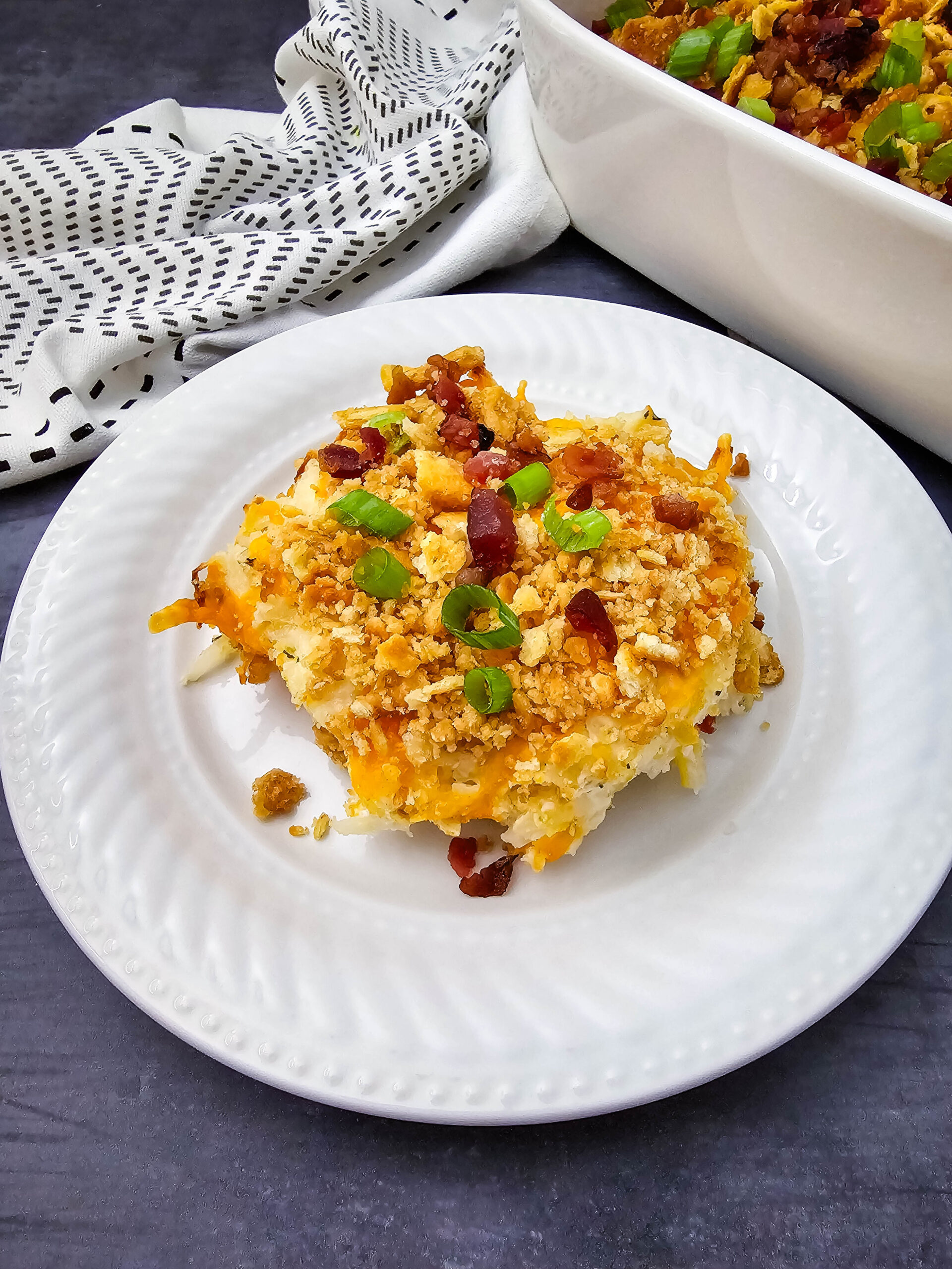 Serving of Million Dollar Potato Casserole on a white plate with buttery cracker topping, bacon pieces, and green onions.