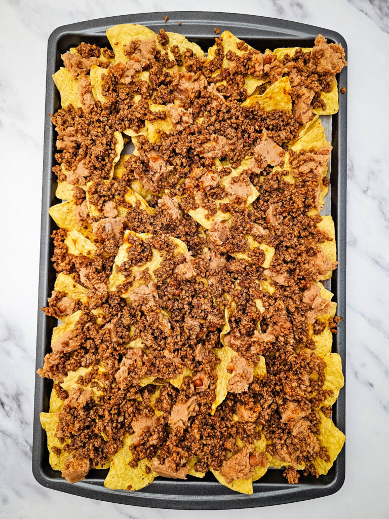 Tortilla chips layered with refried beans and seasoned ground beef on a baking sheet.