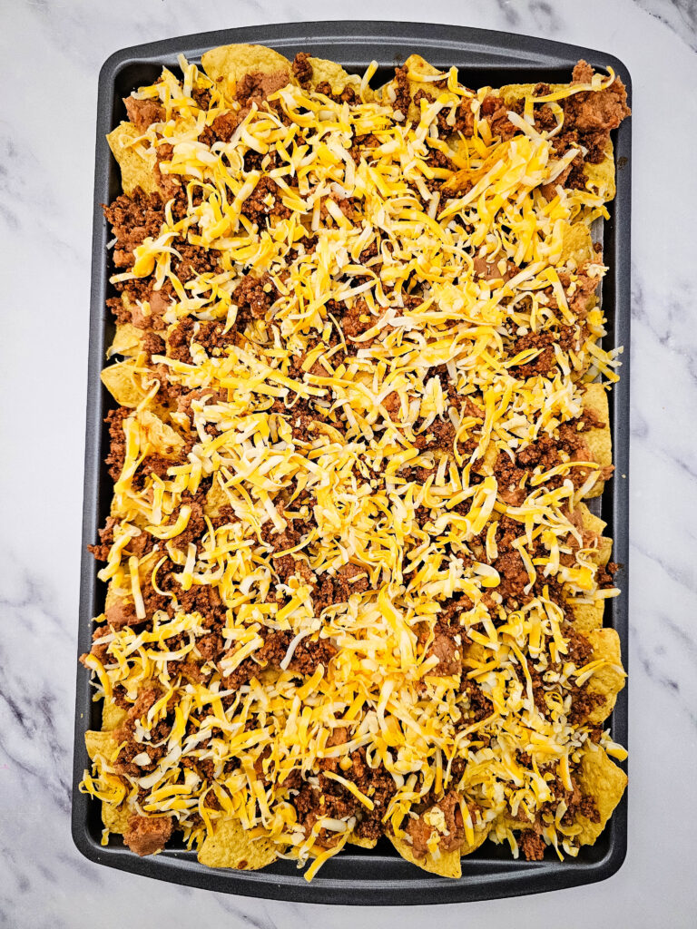 Tortilla chips topped with seasoned ground beef, refried beans, and shredded cheese before baking.