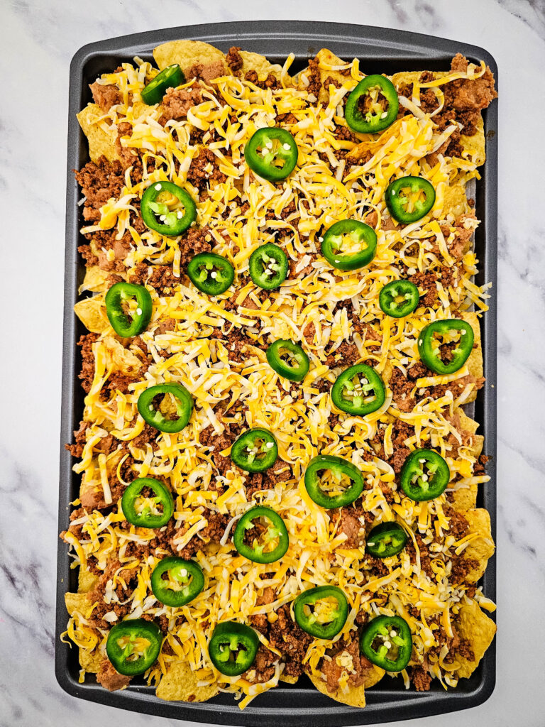 Tortilla chips topped with seasoned ground beef, shredded cheese, and sliced jalapeños before baking.