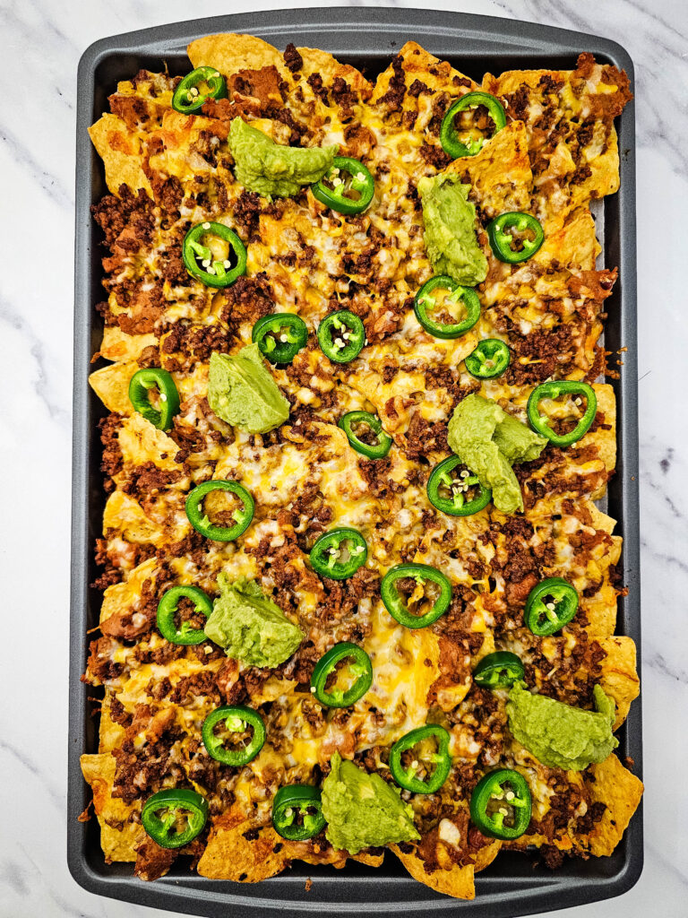 Sheet pan ground beef nachos topped with melted cheese, seasoned beef, jalapeños, and guacamole.