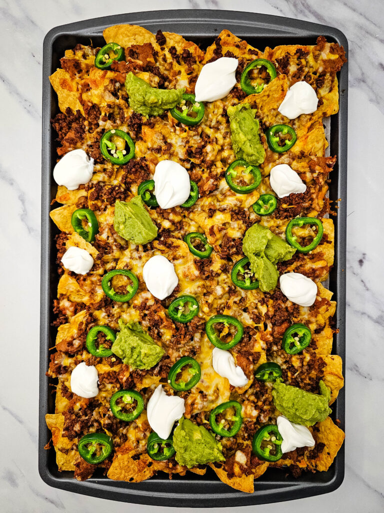Baked sheet pan nachos topped with seasoned beef, melted cheese, jalapeños, sour cream, and guacamole.