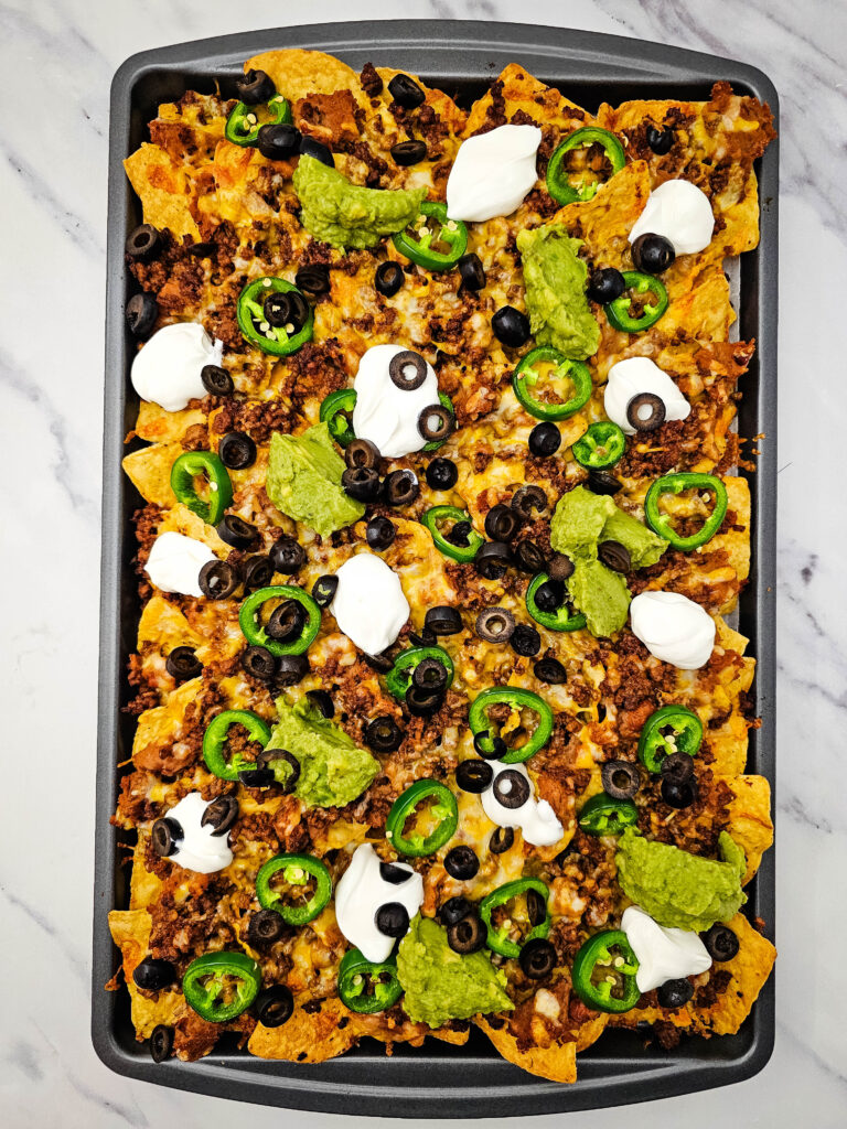 Finished sheet pan ground beef nachos topped with melted cheese, jalapeños, sour cream, guacamole, and black olives.