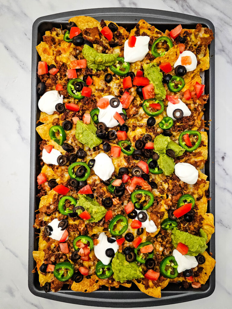 Finished sheet pan ground beef nachos topped with melted cheese, jalapeños, tomatoes, olives, sour cream, and guacamole.
