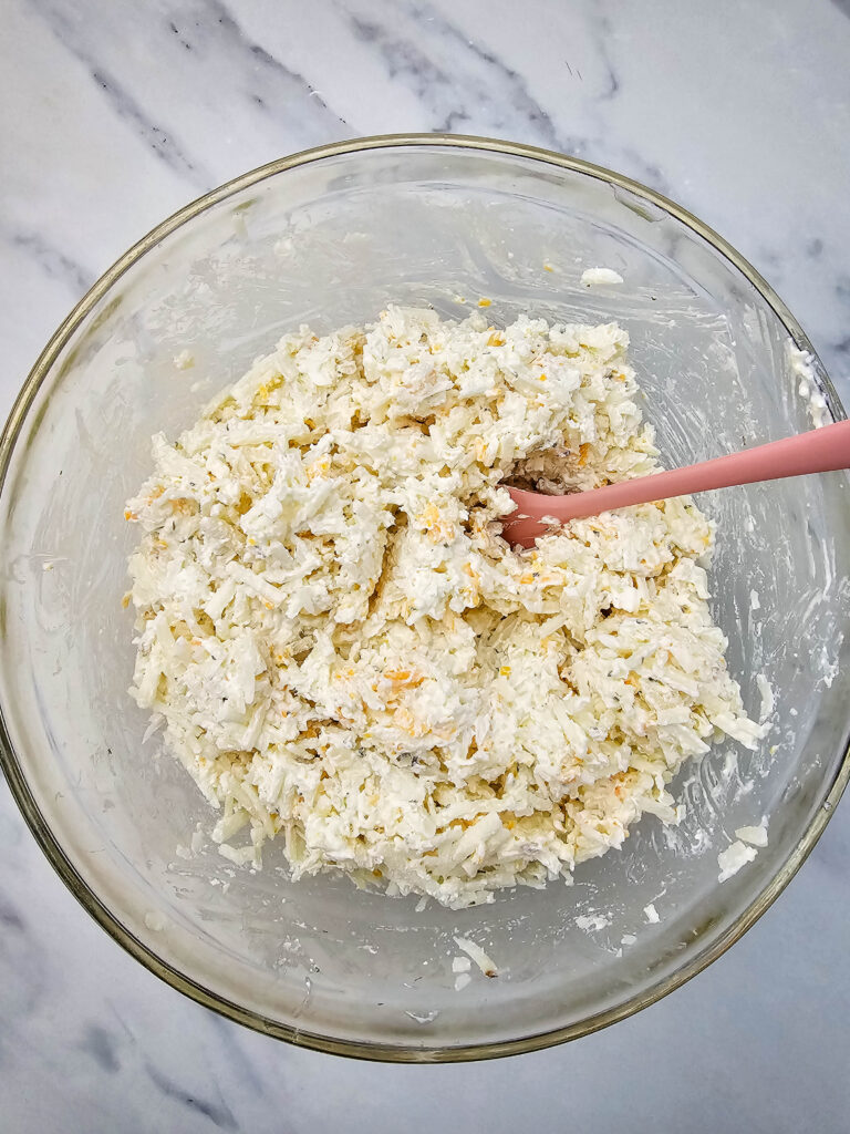 Creamy hashbrown potato mixture in a glass bowl with a pink spatula.