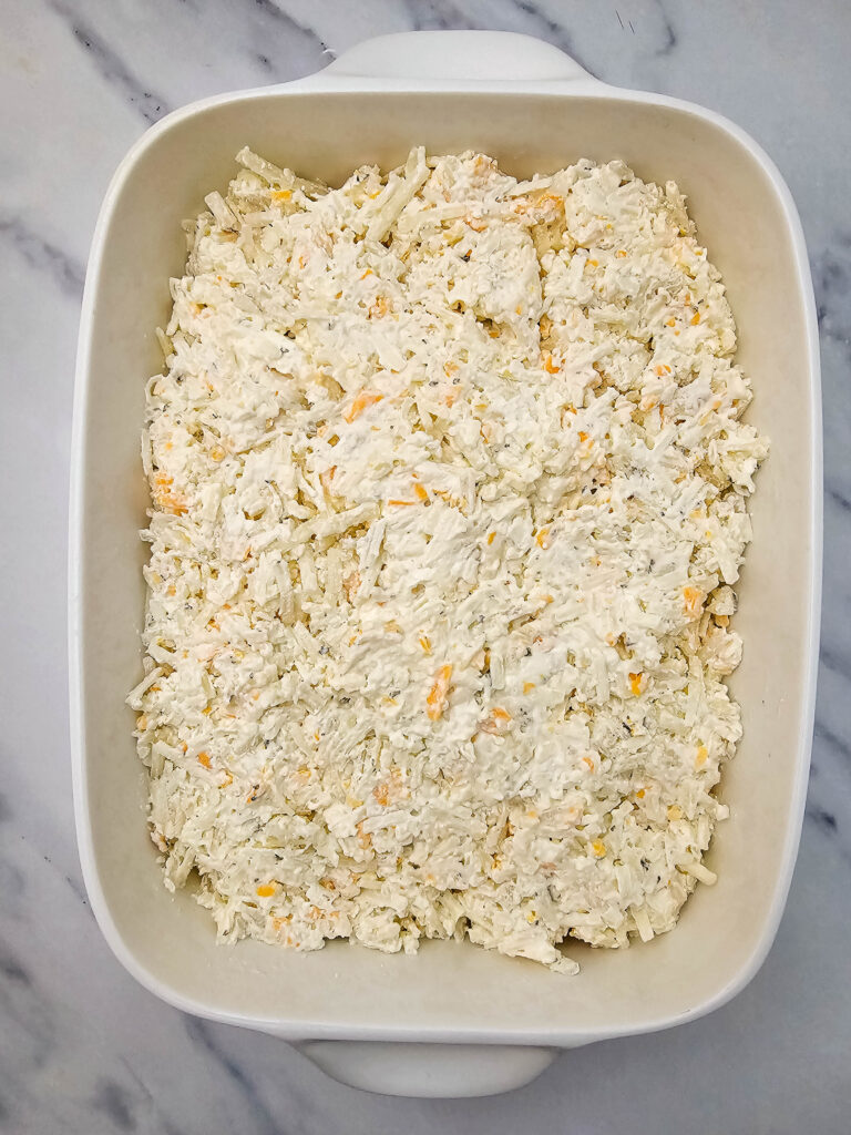 Creamy hashbrown potato mixture spread evenly in a white casserole dish before baking.