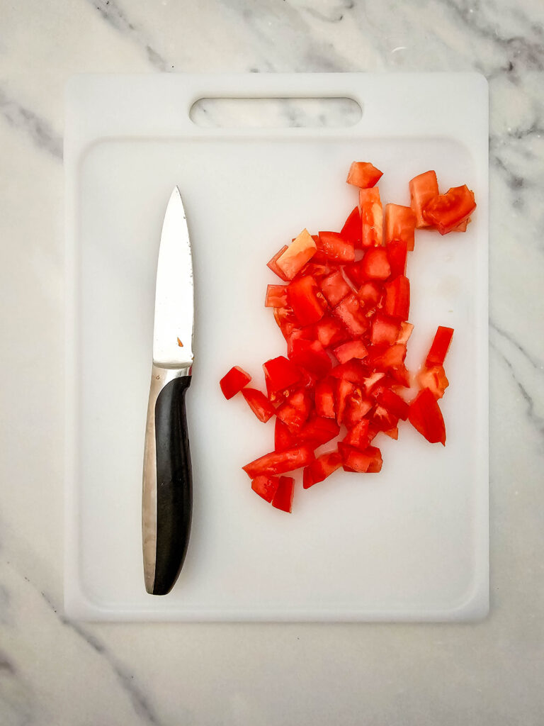 Diced Roma tomato on a white cutting board with a knife for sheet pan ground beef nachos.