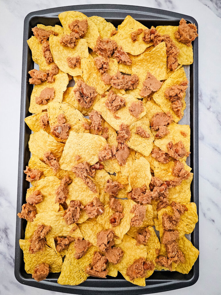 Tortilla chips topped with spoonfuls of refried beans on a baking sheet.
