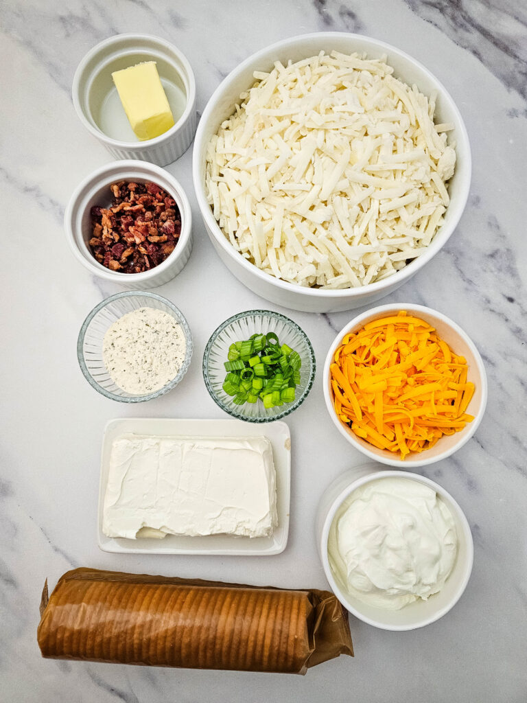 Million Dollar Potato Casserole ingredients arranged on a marble counter, including hashbrowns, cream cheese, sour cream, cheddar cheese, ranch seasoning, bacon, green onions, butter, and crackers.