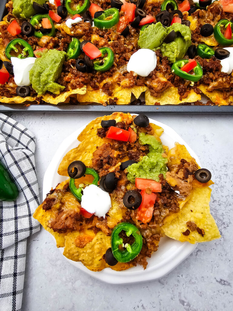 Close-up of loaded ground beef nachos with jalapeños, tomatoes, olives, sour cream, and guacamole served from a sheet pan.