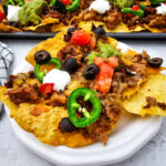 Sheet pan ground beef nachos served on a white plate with jalapeños, tomatoes, olives, sour cream, and guacamole.