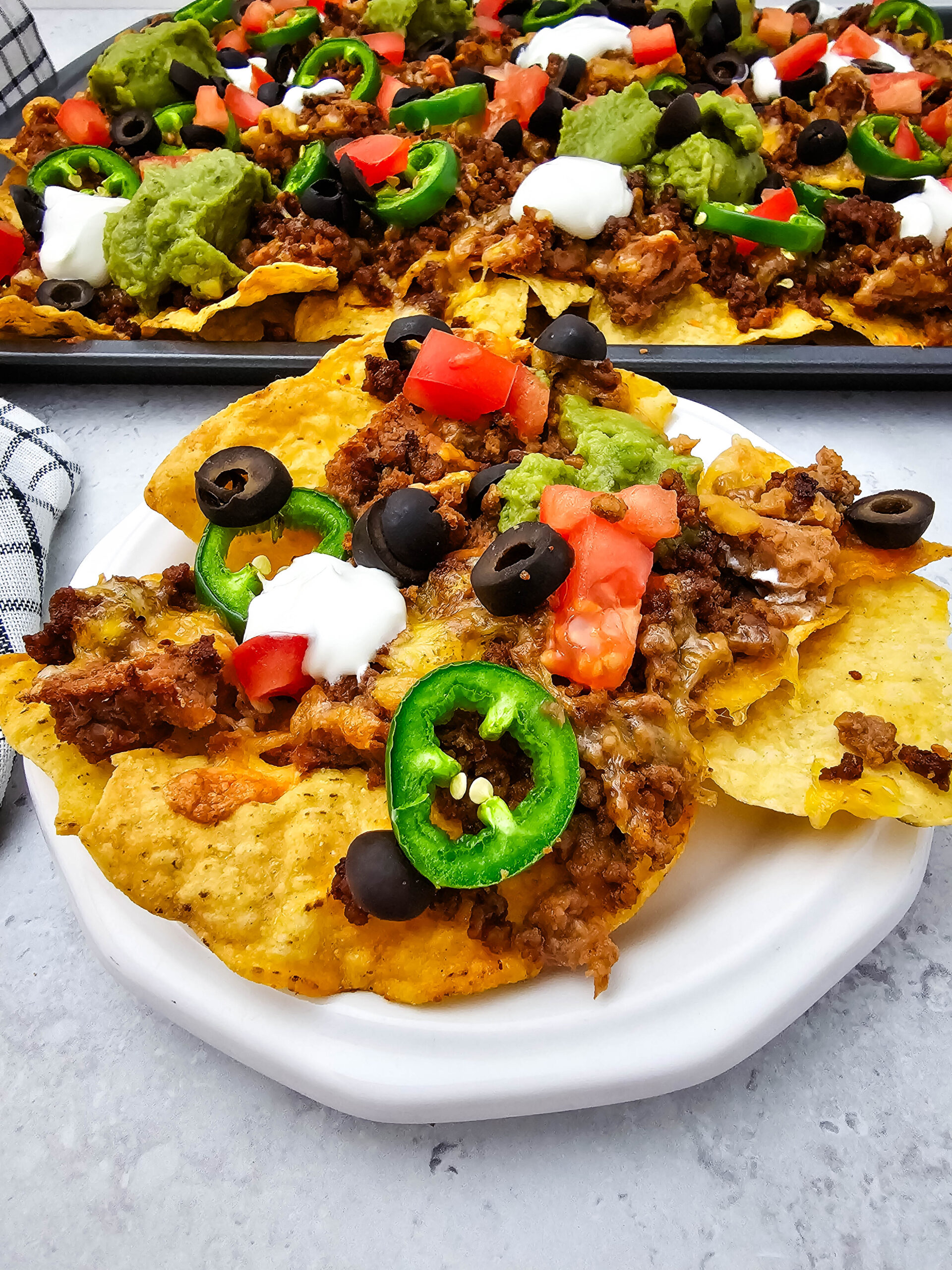 Sheet pan ground beef nachos served on a white plate with jalapeños, tomatoes, olives, sour cream, and guacamole.