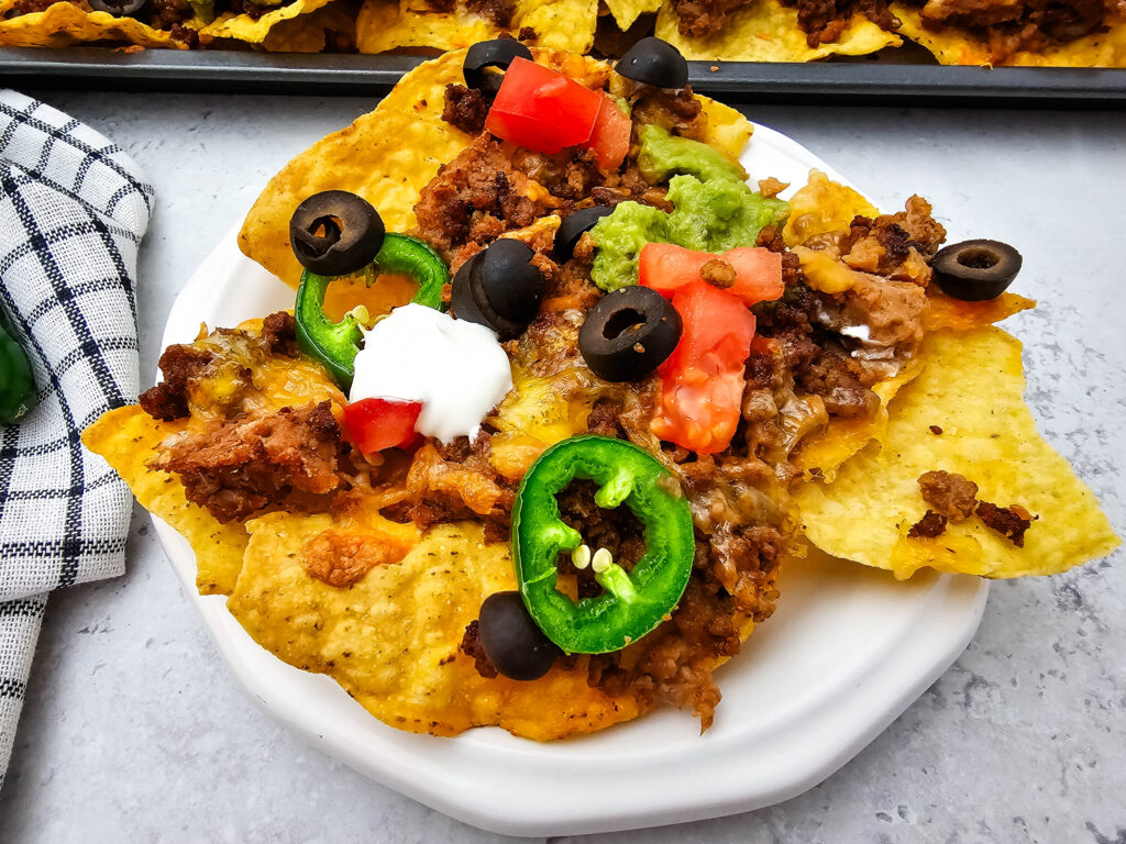 Ground beef nachos piled on a white plate with refried beans, melted cheese, jalapeños, tomatoes, olives, and sour cream.